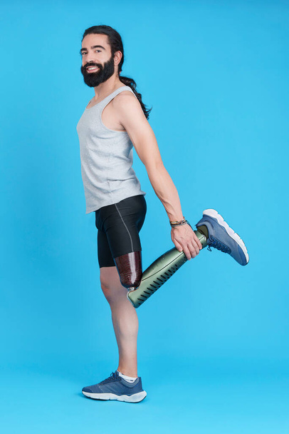 Vertical Studio portrait with blue background of a man with a prosthesis on his leg stretching his upper quadriceps - Foto, Imagem
