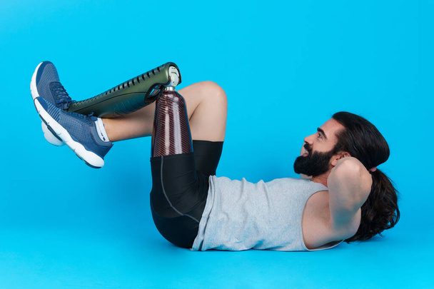Horizontal studio portrait with blue background of a sportive bearded man with a prosthetic leg doing sit-ups - Foto, Imagem