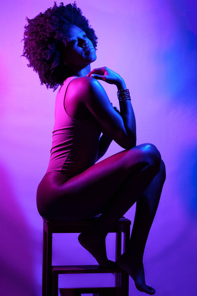Side view of slim African American female model in bodysuit, with curly hair touching shoulder and looking away while sitting on stool under violet neon light - Photo, Image