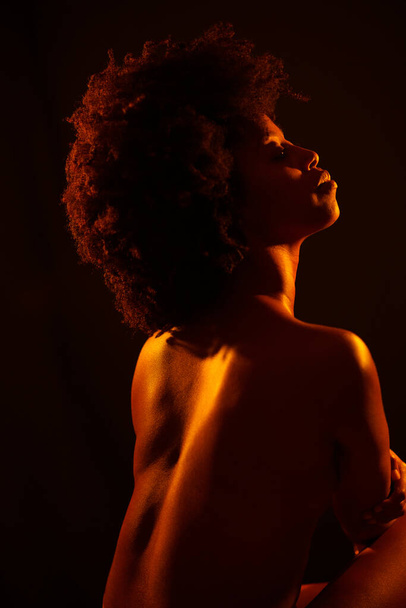 Sensual topless African American female model embracing body and closing eyes under orange neon illumination against black background. - Φωτογραφία, εικόνα