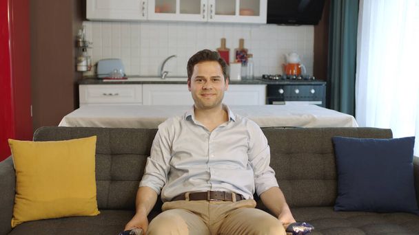 Young man sitting on comfortable sofa in his apartment and looking at camera with smile. Creative people can put whatever they want where the man points. - Foto, immagini