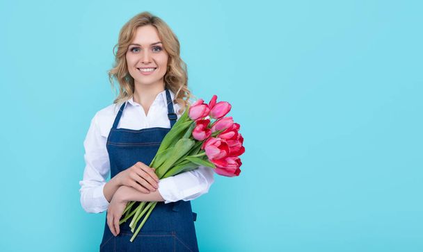 весёлая продавщица цветов в фартуке с весенними цветами тюльпана на синем фоне. - Фото, изображение
