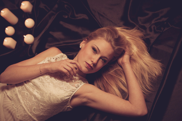 Young blonde woman on black sheets with candels in background - Photo, Image