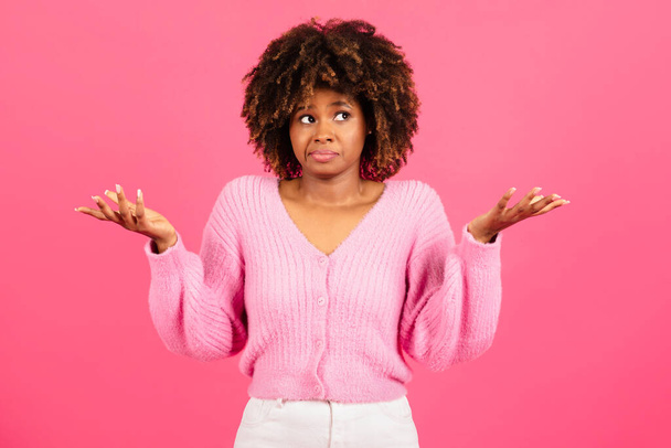 Insecure confused incompetent millennial african american curly lady in casual spreads her arms to sides and holds empty space isolated on pink background, studio. Human emotions, choice, question - Foto, Bild