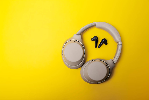 Light gray wireless on-ear headphones and blue in-ear wireless headphones on a yellow background. Comparison of different types of headphones. Headphones for playing games or listening to music. Noise canceling headphones. Top view. Copy space. - Φωτογραφία, εικόνα