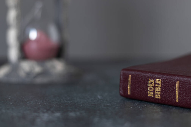 Holy bible book with an hourglass in the background. Copy space for text. Christian biblical concept of time running, final hour, and patience. - Photo, Image