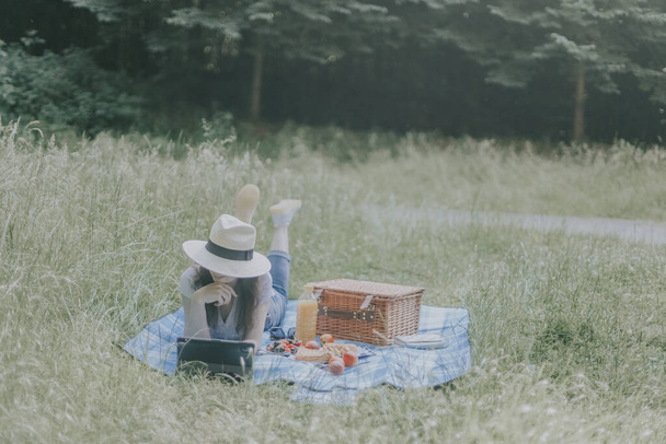 A young beautiful caucasian girl in a straw hat, brown hair, jeans and a t-shirt lies on the floor with a wicker basket, fruits in a plate and browses social networks on a tablet propping her chin - Zdjęcie, obraz
