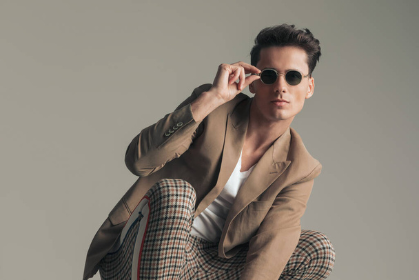 attractive man with dark hair crouching and posing while adjusting sunglasses in front of grey backgroudn in studio - Photo, Image
