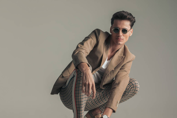 happy man with sunglasses wearing beige jacket and plaid pants and crouching in front of grey background in studio - Foto, immagini