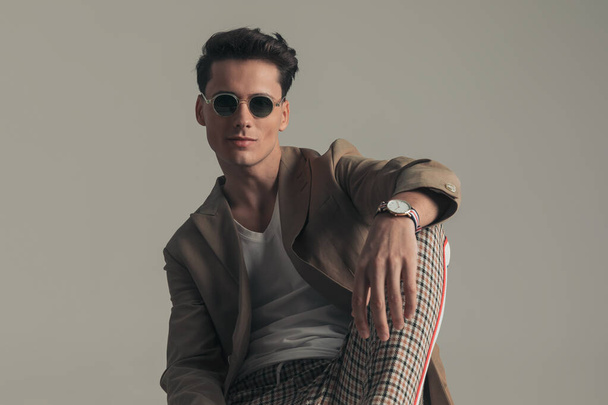 happy confident man wearing sunglasses and beige jacket with plaid pants holding elbow on knee and posing while sitting on grey background - Foto, afbeelding
