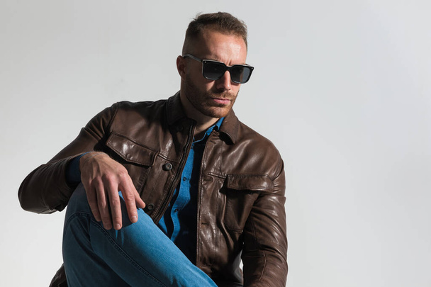 cool fashion man wearing sunglasses, holding knee up with hand above, looking away and posing on grey background in studio - Photo, Image