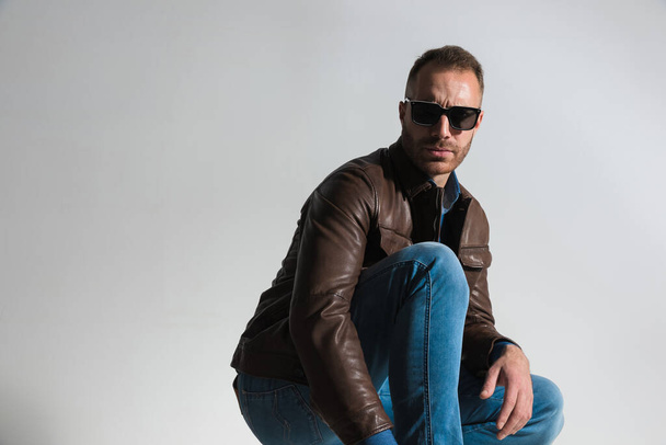 side view picture of sexy confident man in brown leather jacket crouching, wearing brown leather jacket and sunglasses and posing on grey background in studio - Fotografie, Obrázek