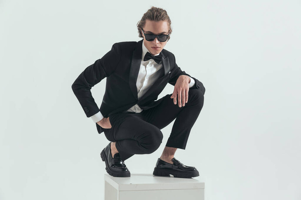 sexy elegant man with blone hair crouching with hand in pockets while posing with the other hand above knee in front of grey background - Фото, изображение