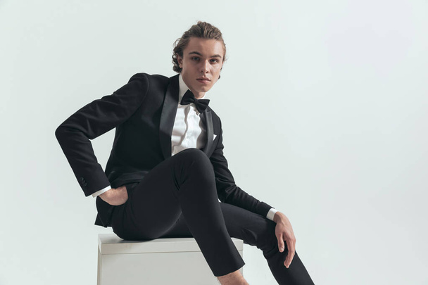 elegant young groom with blue eyes in tuxedo posing with hands in pockets while sitting in front of grey background in studio - Zdjęcie, obraz