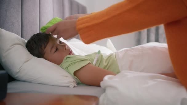 Concerned mother touching her sick little son while he lying in bed with towel on head and measure temperature with thermometer. Preschool kid has fever and coronavirus symptoms concept slow motion - Filmagem, Vídeo