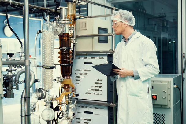 Аптекарь ученый с помощью планшета и ручки для записи информации из экстрактора масла CBD и научной машины, используемой для создания лекарственных продуктов каннабиса. - Фото, изображение