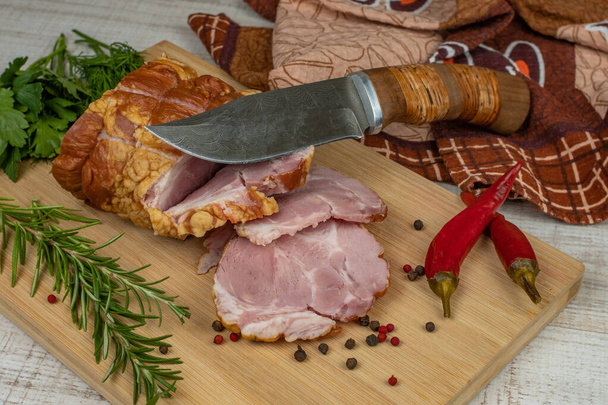 Traditional ham sliced on a wooden cutting board with a carving knife, multi-colored allspice, red hot peppers, parsley, dill and basil. The concept of snacks from pork meat. - Foto, afbeelding