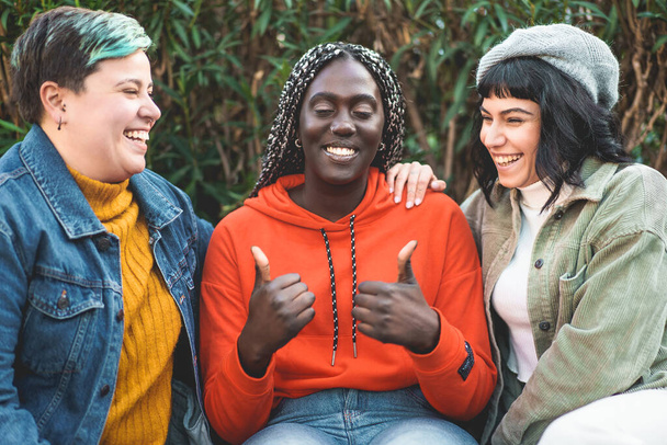 Three young women, an African woman with dreadlocks, a curvy woman with non-binary gender, and a brunette, are seen spending time together chatting and having fun. The African woman is in the center, with both thumbs up, while the other two women hav - 写真・画像