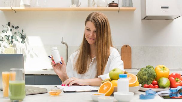 A young nutritionist advises a patient online on taking medications, dietary supplements, showing a jar of vitamins in a laptop camera. Healthy life style. - Photo, Image