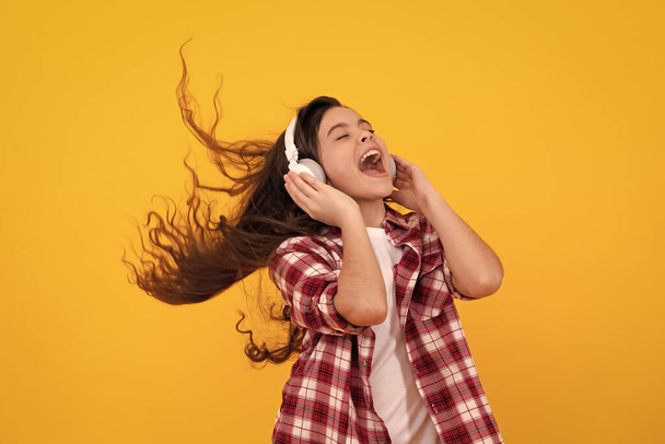 Amazed teenager. Teenager child girl in headphones listening music, wearing stylish casual outfit isolated over yellow background. Excited teen girl - 写真・画像