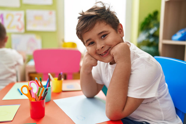 Suloinen latino poika opiskelija hymyilee luottavainen istuu pöydällä päiväkodissa - Valokuva, kuva