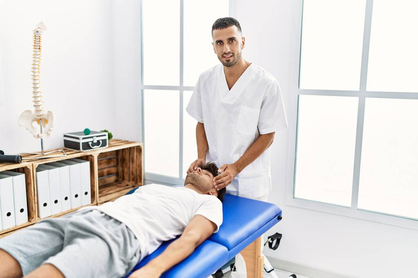 Два испаноязычных мужчины физиотерапевта и пациента, проходящие реабилитацию сеанс массажа головы в клинике - Фото, изображение