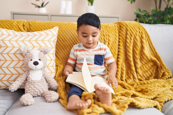 Adorable niño hispano leyendo libro sentado en sofá con osito de peluche en casa - Foto, Imagen