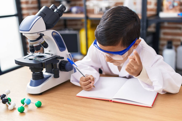 Adorable estudiante hispano usando escritura de microscopio en un cuaderno en el aula de laboratorio - Foto, imagen