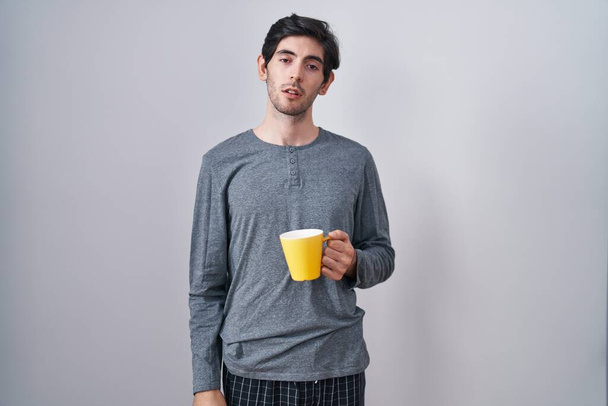 Young hispanic man wearing pajama drinking a cup of coffee looking sleepy and tired, exhausted for fatigue and hangover, lazy eyes in the morning.  - Foto, imagen