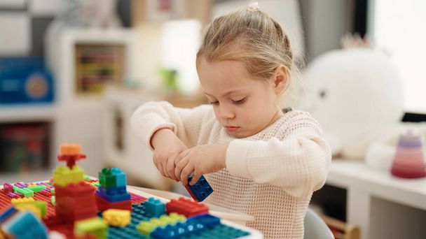 Schattig blond meisje spelen met bouwstenen zitten op tafel op de kleuterschool - Foto, afbeelding