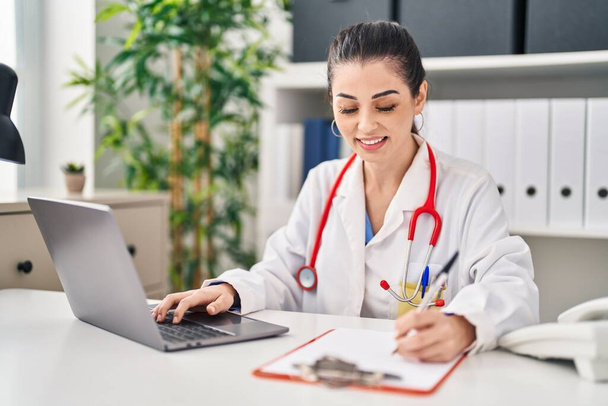 Young beautiful hispanic woman doctor using laptop writing on document at clinic - Photo, Image