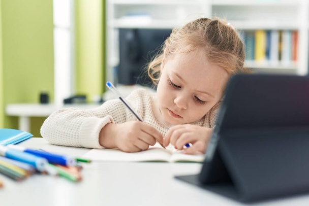 Adorable blonde girl student using touchpad writing on notebook at classroom - Photo, Image