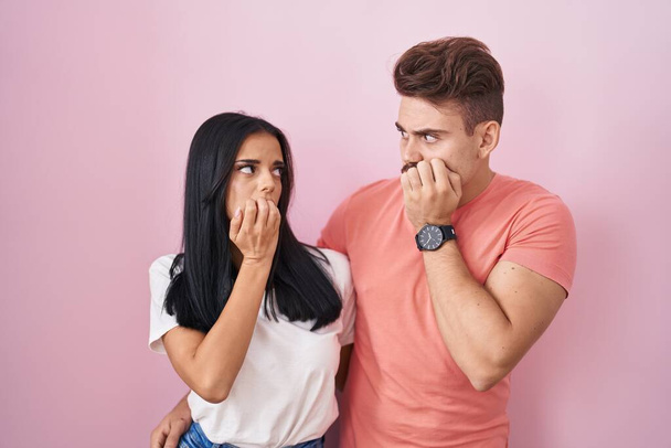 Nuori latino pari seisoo vaaleanpunainen tausta näyttää stressaantunut ja hermostunut kädet suulla puree kynnet. ahdistuneisuusongelma.  - Valokuva, kuva