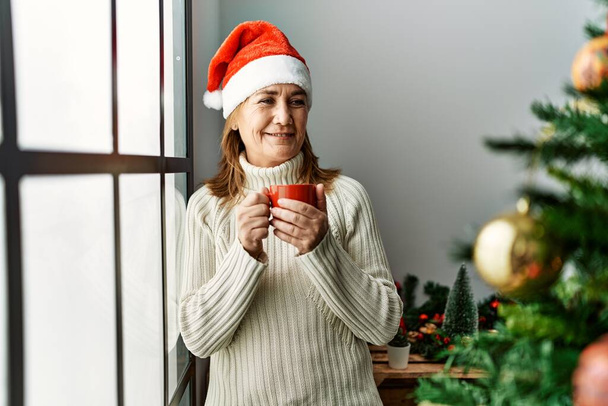 Orta yaşlı beyaz kadın evde Noel ağacının yanında kahve içiyor. - Fotoğraf, Görsel