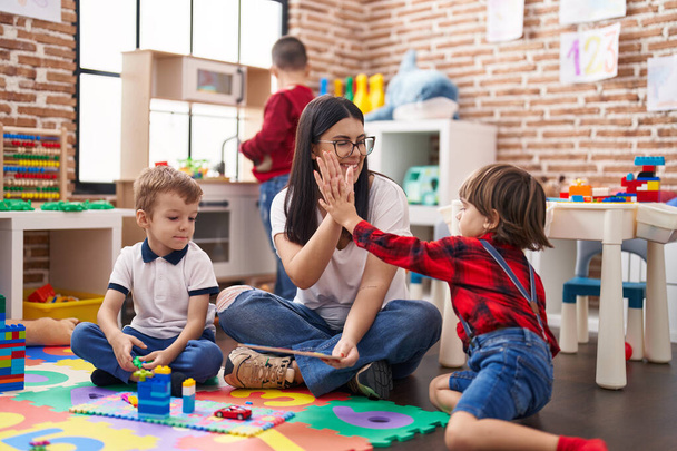 Lehrerin mit einer Gruppe von Jungen, die mit erhobenen Händen im Kindergarten auf dem Boden hoch fünf sitzt - Foto, Bild