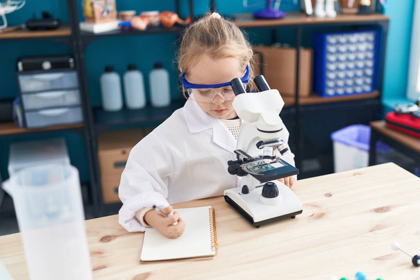 Adorable blonde girl student using microscope writing on notebook at laboratory classroom - 写真・画像