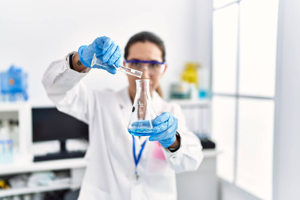 Νεαρή ισπανόφωνη γυναίκα με στολή επιστήμονα που κρατά δοκιμαστικό σωλήνα στο εργαστήριο - Φωτογραφία, εικόνα