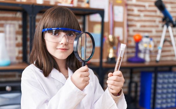 Adorable hispanic girl student looking test tube using magnifying glass at laboratory classroom - Foto, imagen