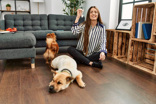 Young hispanic woman holding key of new house sitting on floor with dogs at home - Foto, imagen