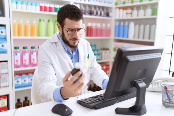 Nuori latino mies farmaseutti käyttäen tietokoneen tilalla lääkitys pullo apteekissa - Valokuva, kuva