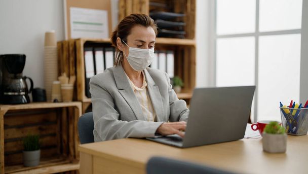 Середнього віку його іспанська жінка бізнесмен в медичній масці працює в офісі
 - Фото, зображення