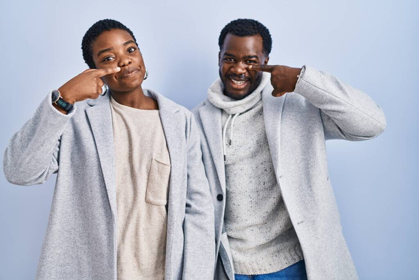 Νεαρό ζευγάρι αφροαμερικανών στέκεται πάνω από το μπλε φόντο μαζί δείχνοντας με το χέρι το δάχτυλο στο πρόσωπο και τη μύτη, χαμογελώντας χαρούμενα. έννοια ομορφιά  - Φωτογραφία, εικόνα
