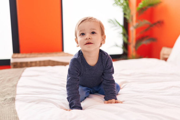 Imádnivaló szőke kisbaba ül az ágyon nyugodt arckifejezéssel a hálószobában - Fotó, kép