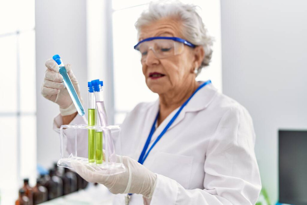 Senior grey-haired woman wearing scientist uniform holding test tubes at laboratory - Фото, изображение