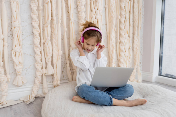 Valkoihoinen hauska lapsi kuulokkeissa, istuu tietokoneen kanssa, kannettava tietokone kotona. Koulutyttö, oppilas pitää hauskaa, käyttää gadgetia, kuunnella musiikkia, katsoa videoluokkaa, pelata videopelejä. Nuori bloggaaja. - Valokuva, kuva