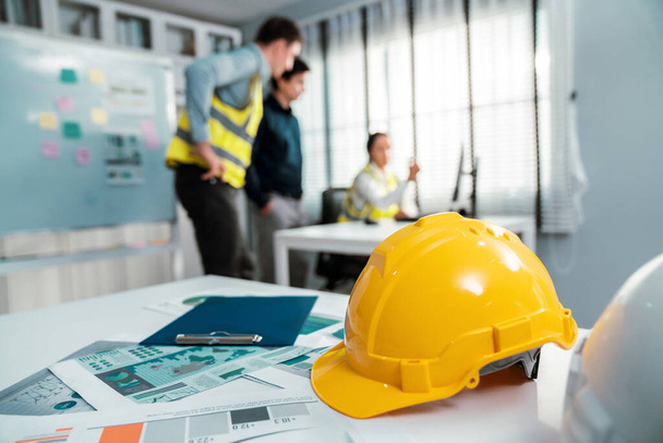 Concept of engineering equipments of protective helmet for competent engineer on piles of paper works. Blurred image of engineers and employer working together at the desk. - Valokuva, kuva