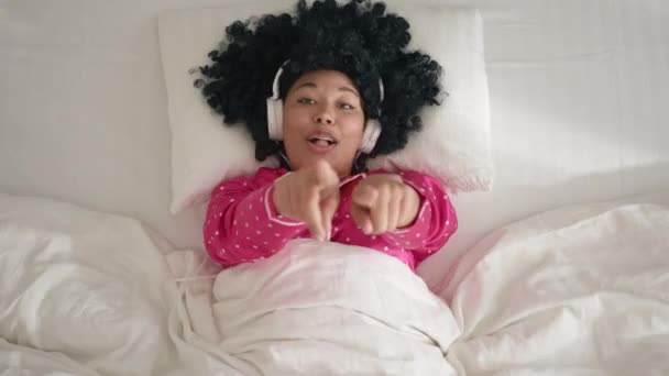 Joyful girl with afro hairstyle is dancing hands and singing wearing headphones having fun on bed at home. Modern entertainment, music and happy young people concept, slow motion funny woman of color - Video, Çekim
