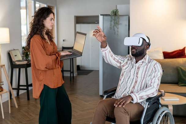 Young woman with laptop standing in front of man with disability sitting in wheelchair with vr headset and pointing at virtual screen - Foto, imagen
