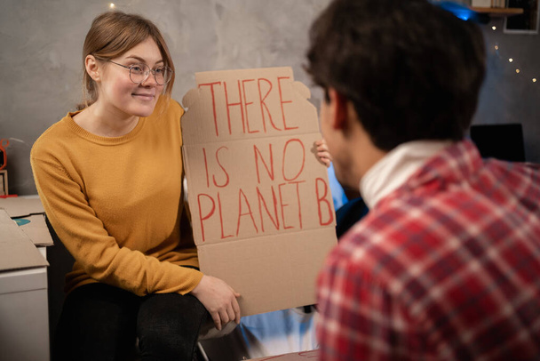 Students prepare banner protesting over pollution and global warming sitting in dormitory. Protest to save planet earth. Woman showing a banner of There is no Planet B for her boyfriend. Close up - Foto, immagini