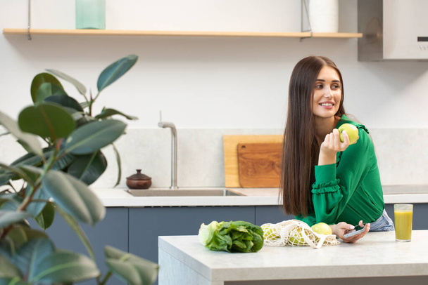 An attractive brunette holds a green apple and smiles while standing at the kitchen table. Healthy lifestyle - Foto, immagini